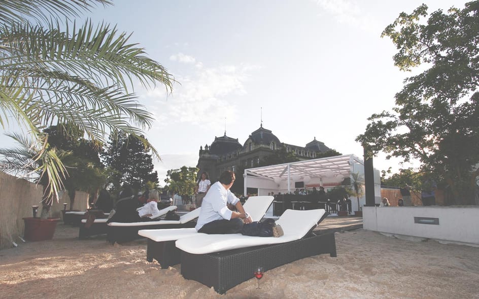 Schwarze Rattanliegen mit weissen Auflagen auf einer Sandfläche, dahinter eine Bar unter einer weissen Pergola und ein Kuppelbau aus dem 19. Jahrhundert