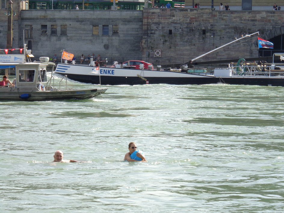 Zwei Schwimmende treiben im Rhein bei Basel, eine mit blauem Schwimmsack; dahinter ein Frachtschiff am Quai und darüber ein Tram auf der Uferstrasse