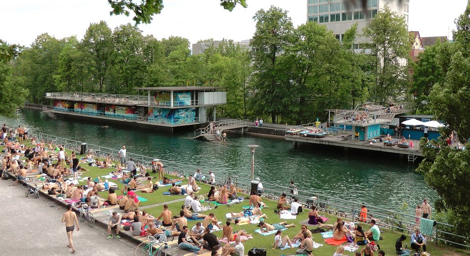 Das Flussbad Oberer Letten an der Limmat in Zürich: volle Liegewiese, Holzstege und der Badeturm am Wasser